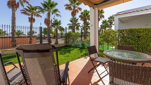 A covered outdoor seating area with wicker furniture, a glass table, and a view of palm trees and a fence.