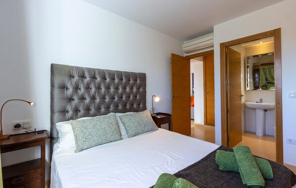 Un dormitorio con un gran cabecero capitonado, una cama con almohadas estampadas, una mesita de noche con lámpara y una toalla verde sobre la cama.