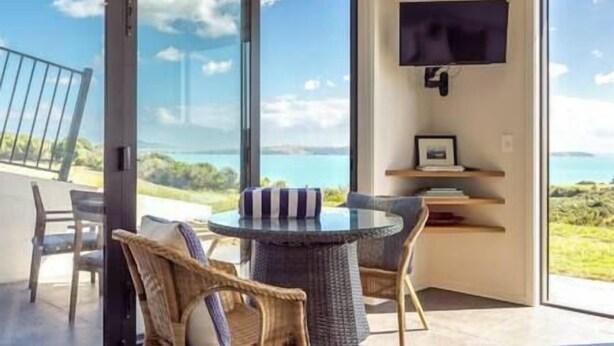 Une chambre moderne avec une porte vitrée donnant sur un balcon qui offre une vue sur un plan d’eau. On y trouve une table ronde avec des chaises, un téléviseur à écran plat fixé au mur, ainsi qu’une petite étagère avec des livres et un tableau encadré.