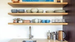 Une cuisine avec des étagères en bois où sont rangés des bols, des assiettes et des tasses, un évier et une bouilloire.