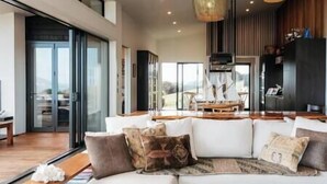 A modern living room with a white sectional sofa, a wooden coffee table, and a large window offering a view of the outdoors. - Tradewinds Estate by Waiheke Pure - Commanding views, pool, hot tub, grand open planned living, sun soaked decks (Waiheke Island)