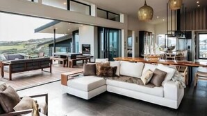 A spacious living area with a large white sectional sofa, a dining table, and a kitchen area with modern appliances. - Tradewinds Estate by Waiheke Pure - Commanding views, pool, hot tub, grand open planned living, sun soaked decks (Waiheke Island)