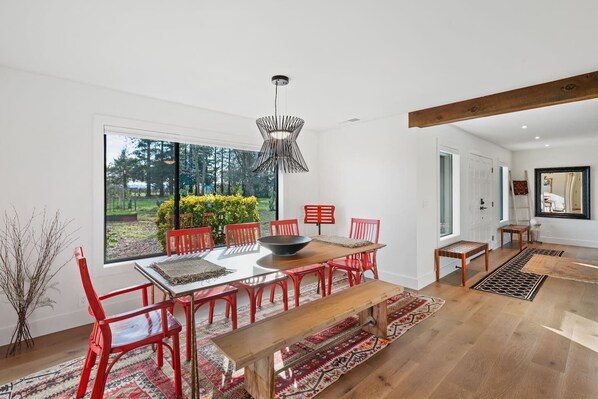 A dining area with a large table, red chairs, a hanging light fixture, and a view of greenery outside.