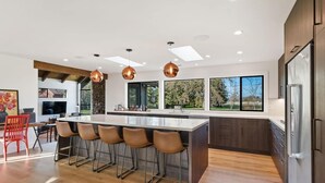A modern kitchen with a central island, bar stools, pendant lighting, and a large window offering a view of the outdoors.