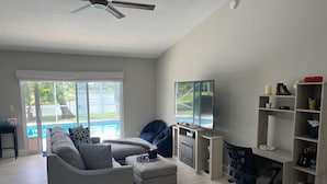 A spacious living room with a grey sectional sofa, a small ottoman, a TV on a wooden stand, and a bookshelf.