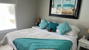 A neatly made bed with a teal throw blanket, a nightstand with a lamp and a clock, and a framed picture of a beach scene on the wall.