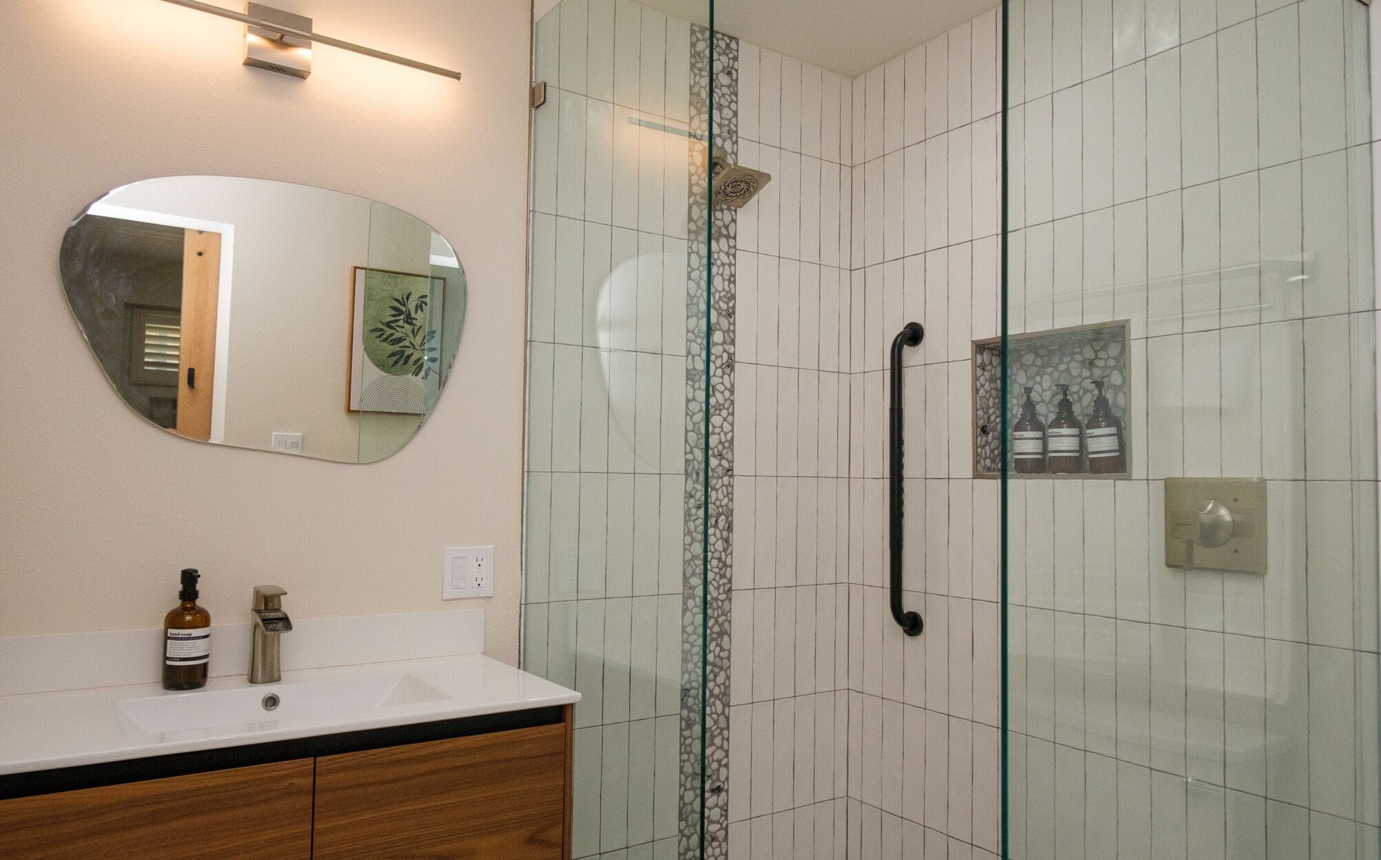 Een badkamer met een douchecabine van glas, een ronde spiegel, een wastafel met houten kastje en een handzeepfles.