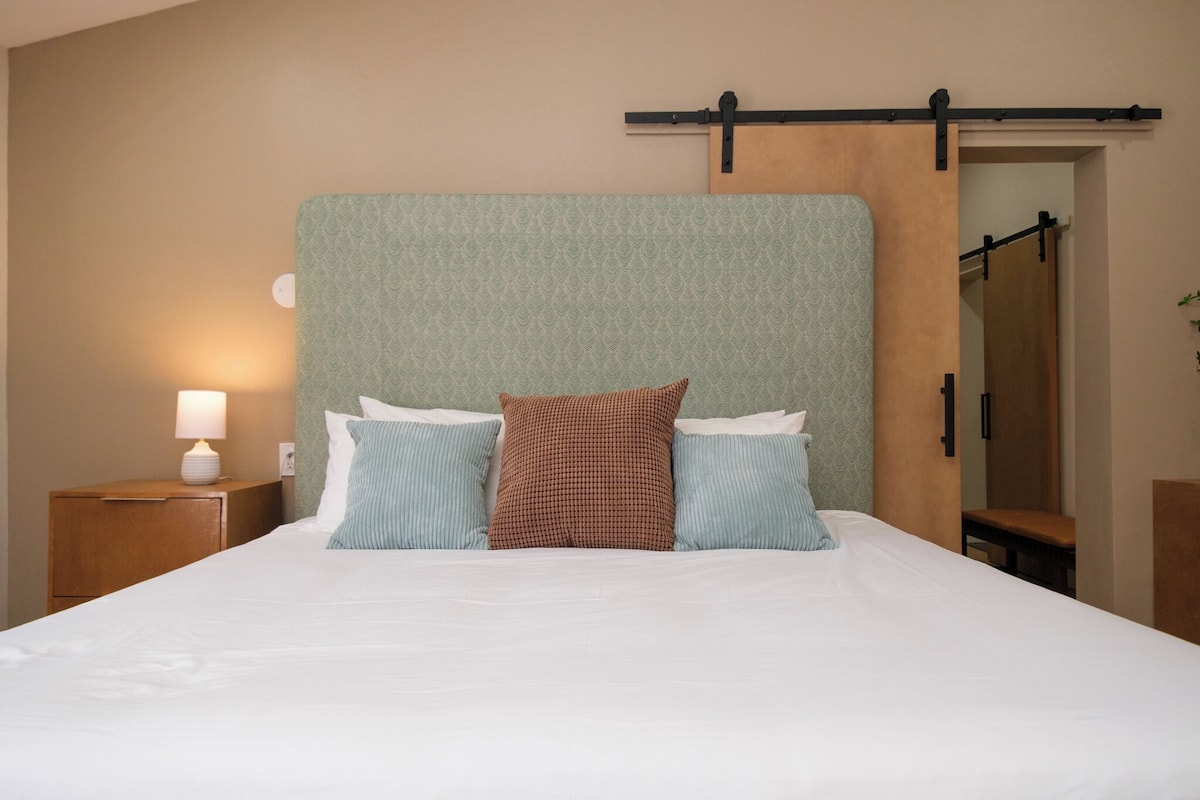 Una cama bien tendida con cabecera, una mesita de noche con lámpara, y un banco de madera.