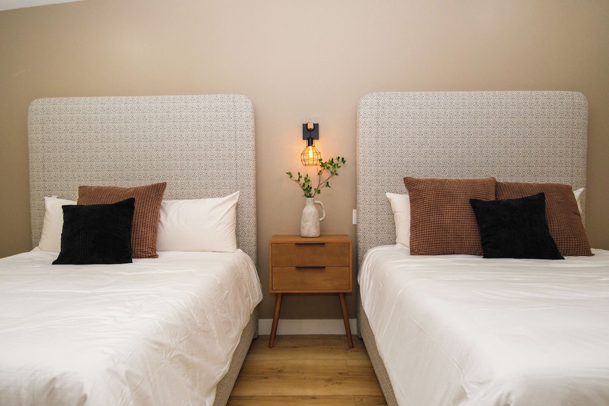 Una habitación de hotel con dos camas, cada una con cabecero estampado, una mesita de noche de madera con un jarrón y una lámpara de pared, y suelo de madera.