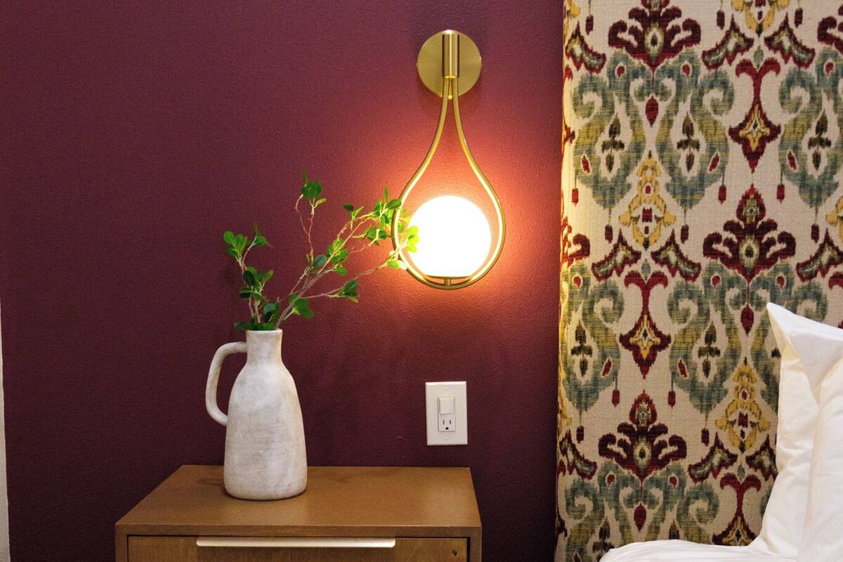 Un dormitorio con una mesita de noche de madera, un jarrón blanco con una planta, un cabecero estampado y un aplique de pared.