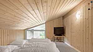 A bedroom with a wooden sloped ceiling, a bed with white bedding, a television on a stand, and a window providing natural light. - Gorgeous home in Væggerløse with sauna (Væggerløse)