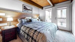 Une chambre avec un plafond en bois, un grand lit recouvert d’une couverture à carreaux, deux tables de chevet avec des lampes et une fenêtre donnant sur l’extérieur.