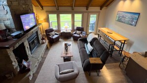 Un salon chaleureux avec une cheminée en pierre, des fauteuils confortables, un plafond en bois et de grandes fenêtres.