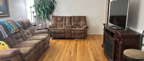 A living room with a brown sofa, a recliner, a TV on a wooden cabinet, and a large plant.