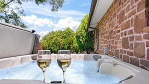 A hot tub with two glasses of white wine, surrounded by a brick wall and greenery.