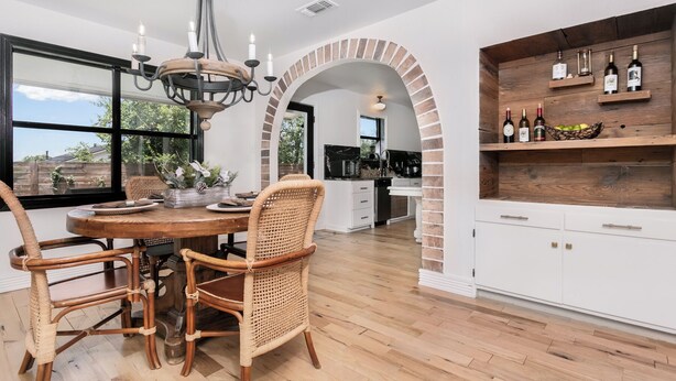 A modern kitchen with a dining area, featuring a brick archway, white cabinetry, and a wine shelf.