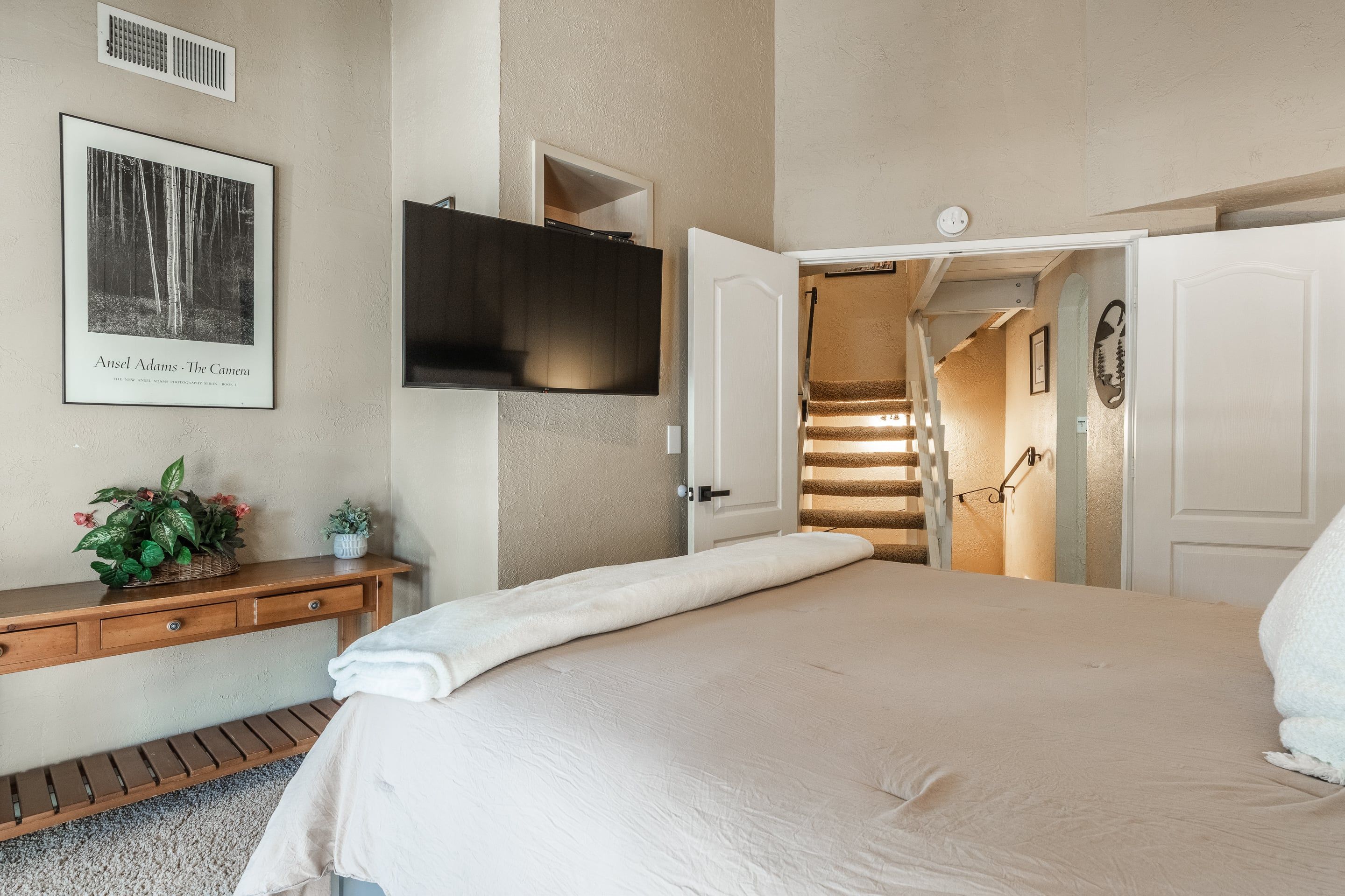 A bedroom with a bed, a flat-screen TV mounted on the wall, a wooden nightstand with a plant, and a staircase leading to another room.