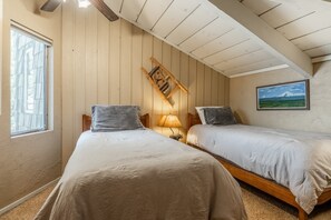 A room with two beds, a wooden slat ceiling, a window, a lamp, a clock, and a framed picture on the wall.