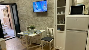 Una cocina pequeña con un refrigerador blanco, un microondas encima y una mesa de comedor con sillas. También hay un televisor montado en la pared.