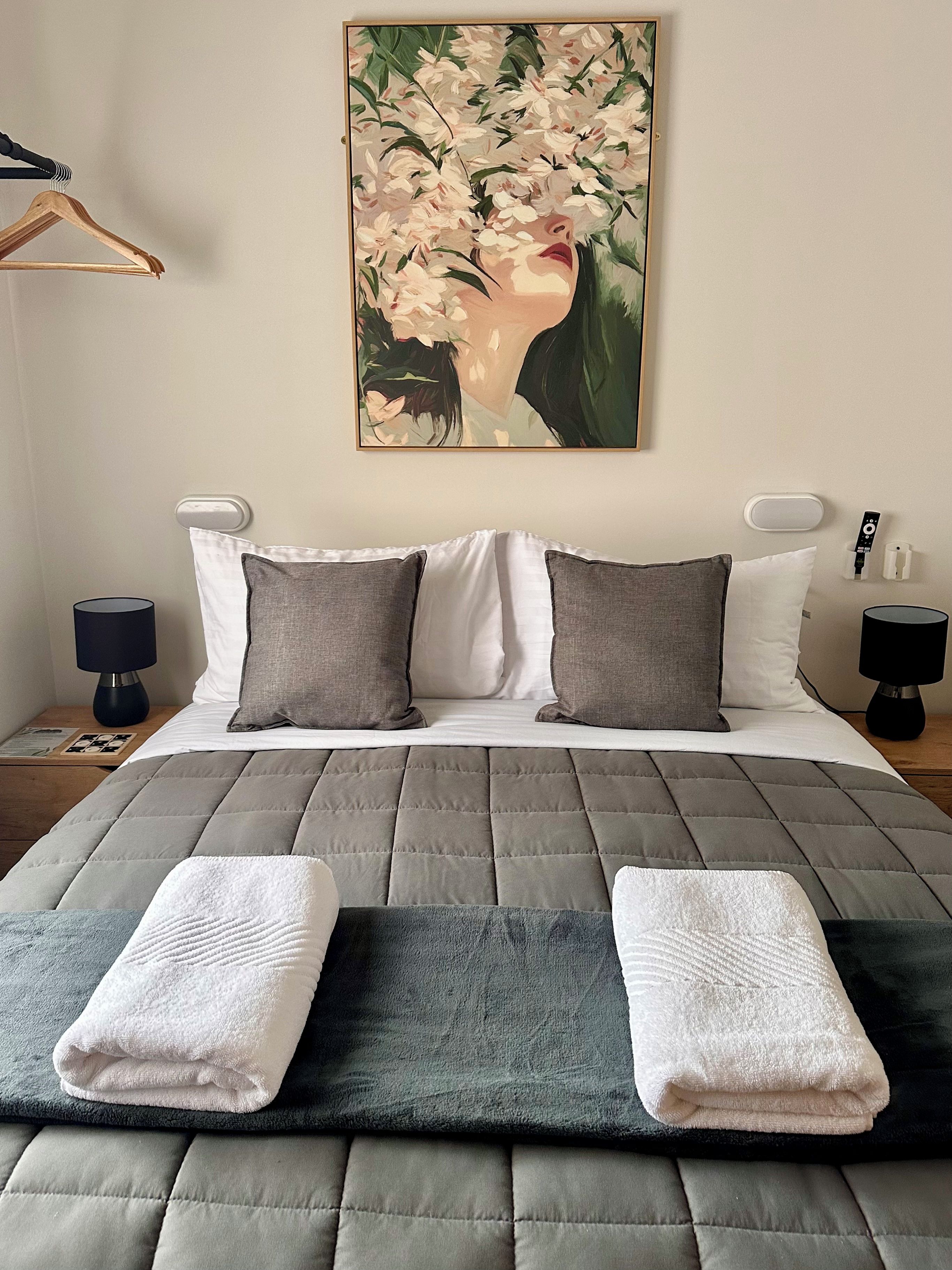 Un dormitorio con cama, almohadas, manta, un cuadro en la pared, lámparas de noche y una mesita.