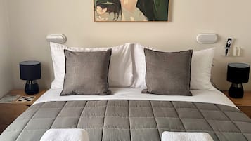 Un dormitorio con cama, almohadas, manta, un cuadro en la pared, lámparas de noche y una mesita.