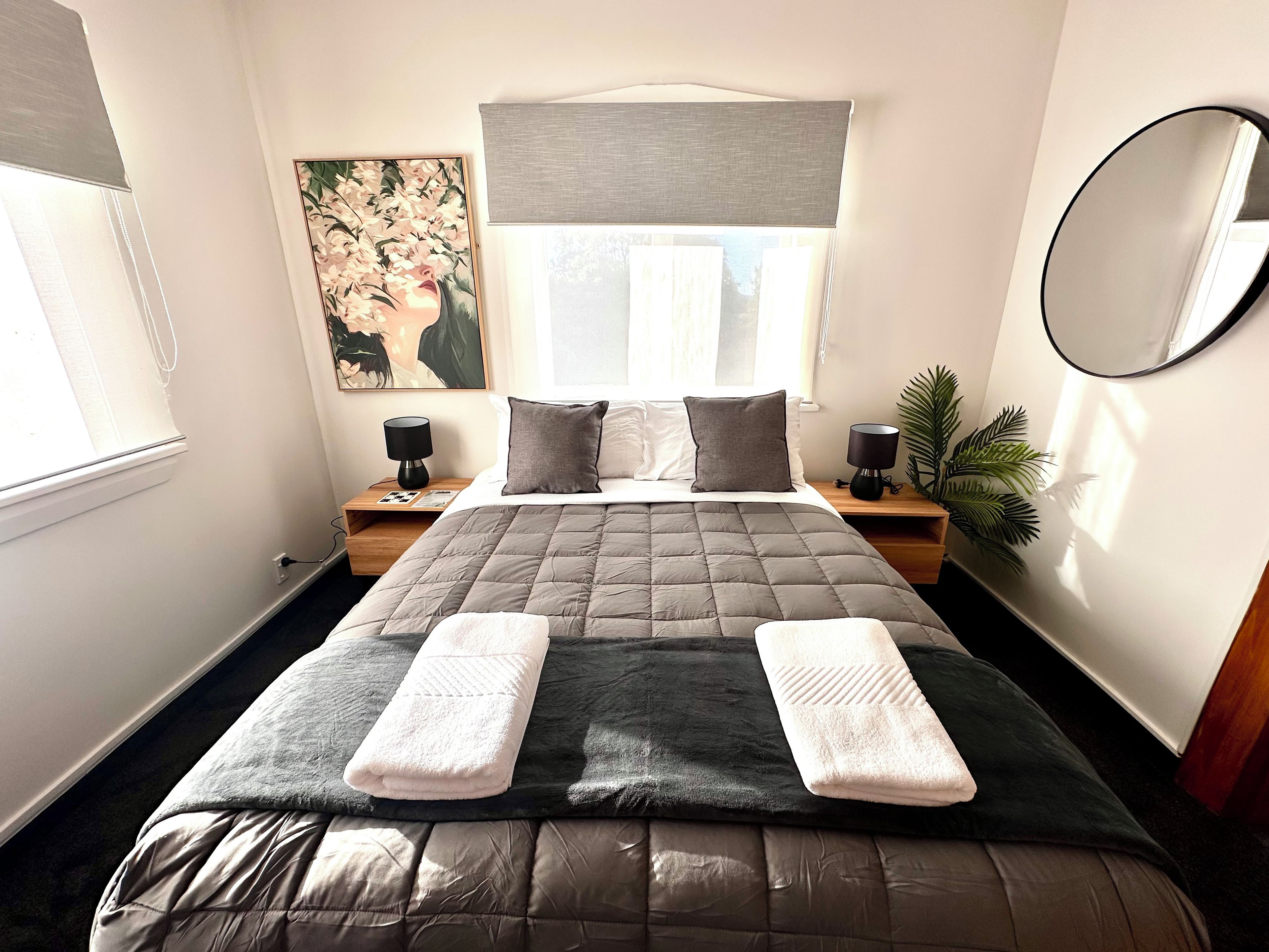 A neatly made bed with a quilted headboard, two bedside lamps, a large mirror, and a painting on the wall.