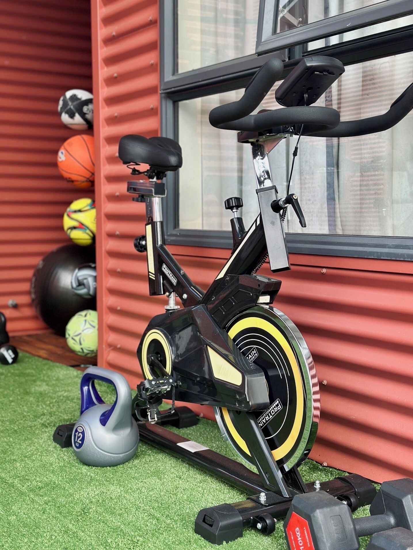 A stationary bike with a water bottle holder, a set of dumbbells, and exercise balls against a red wall.