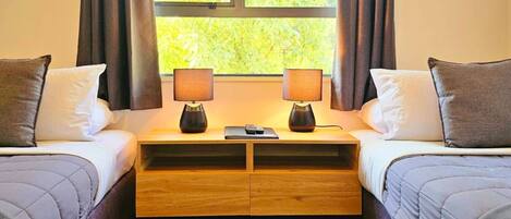 Una habitación de hotel con dos camas, una mesita de noche de madera, dos lámparas y una ventana con cortinas.