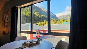 A room with a large window offering a view of a mountainous landscape and a red building. - Wild Kea Lodge Franz Josef (Franz Josef Glacier)