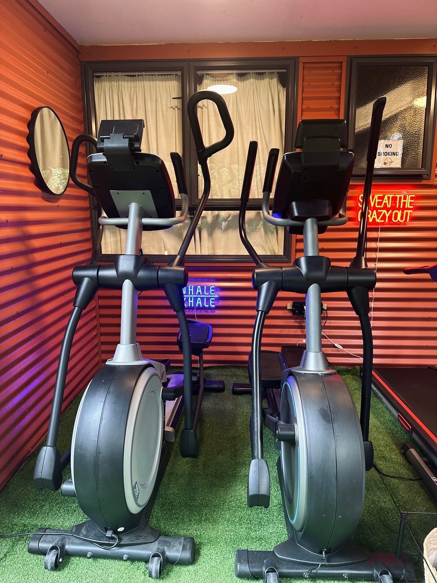 A gym with two elliptical machines, a stepper, and a neon sign that reads "Seattle Crazy Out".