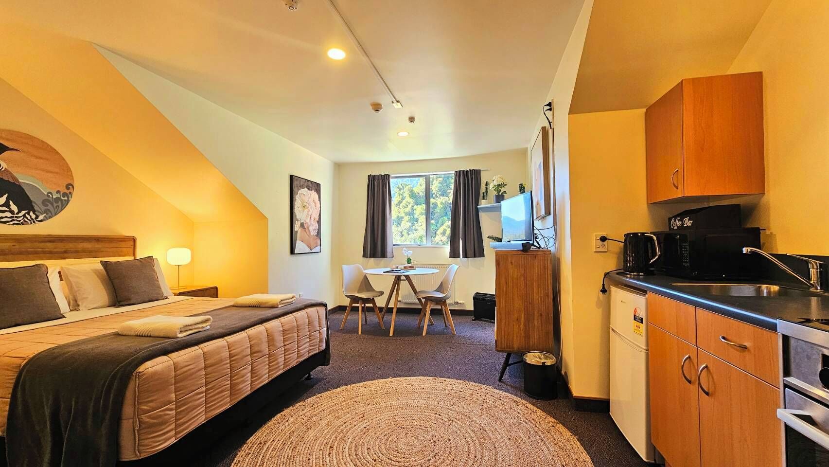 A compact hotel room with a bed, a small desk, a coffee machine, and a kitchenette.