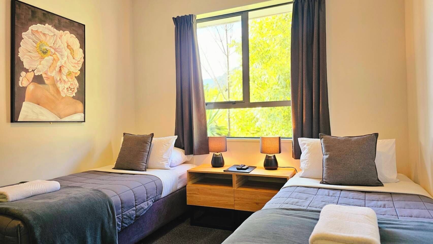 Une chambre d’hôtel avec deux lits simples, une table de chevet avec des lampes, un tableau au mur et une fenêtre avec des rideaux.