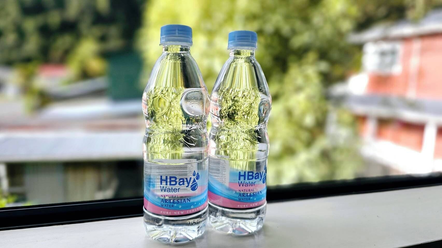 Deux bouteilles d’eau HBay sur un rebord de fenêtre, avec un arrière-plan flou de bâtiments et de verdure.