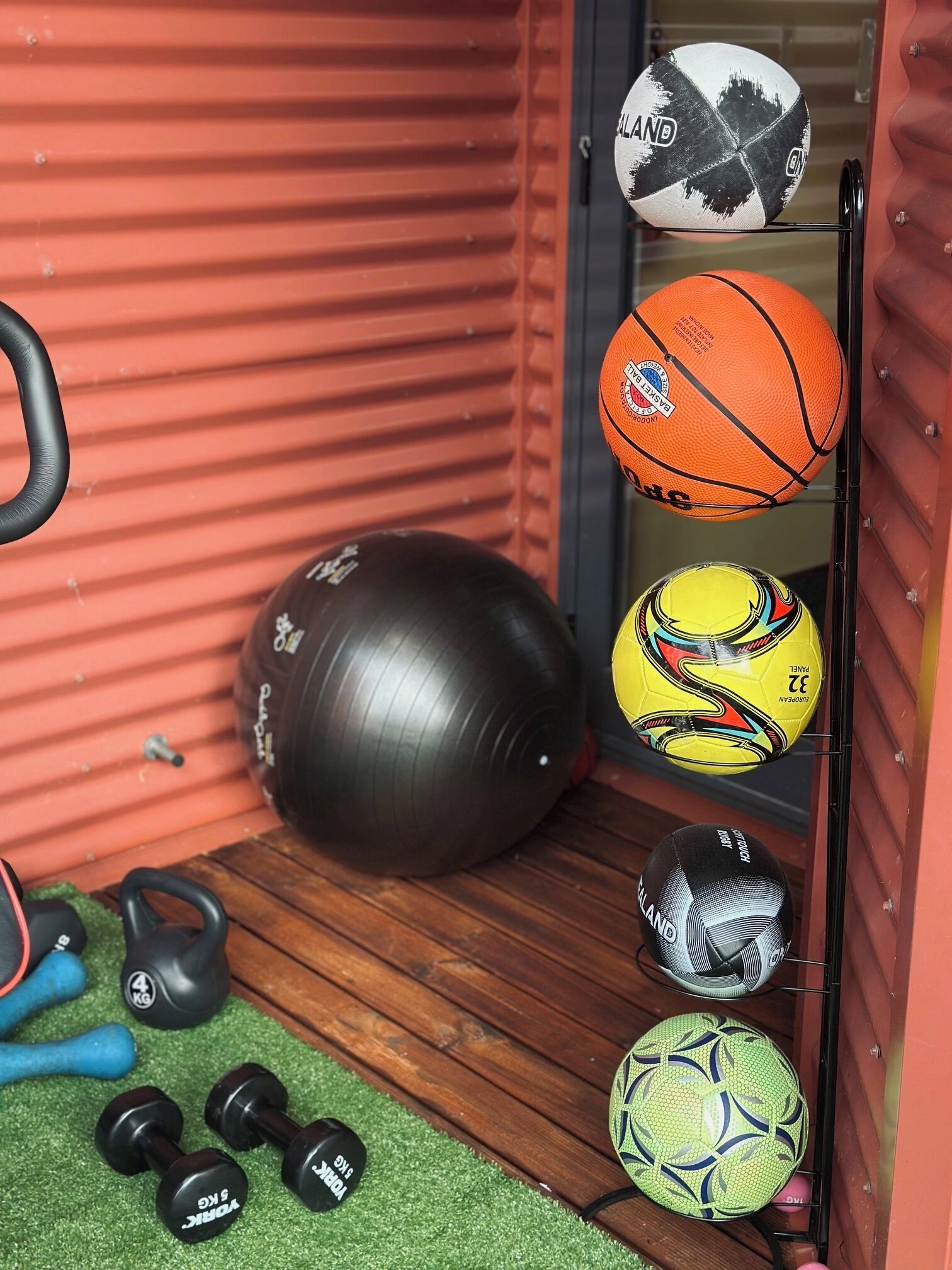 Un área de almacenamiento con equipo deportivo, incluyendo una pelota de ejercicios negra, un balón de fútbol amarillo y negro, un balón de baloncesto blanco y negro, y un balón de fútbol verde y negro.