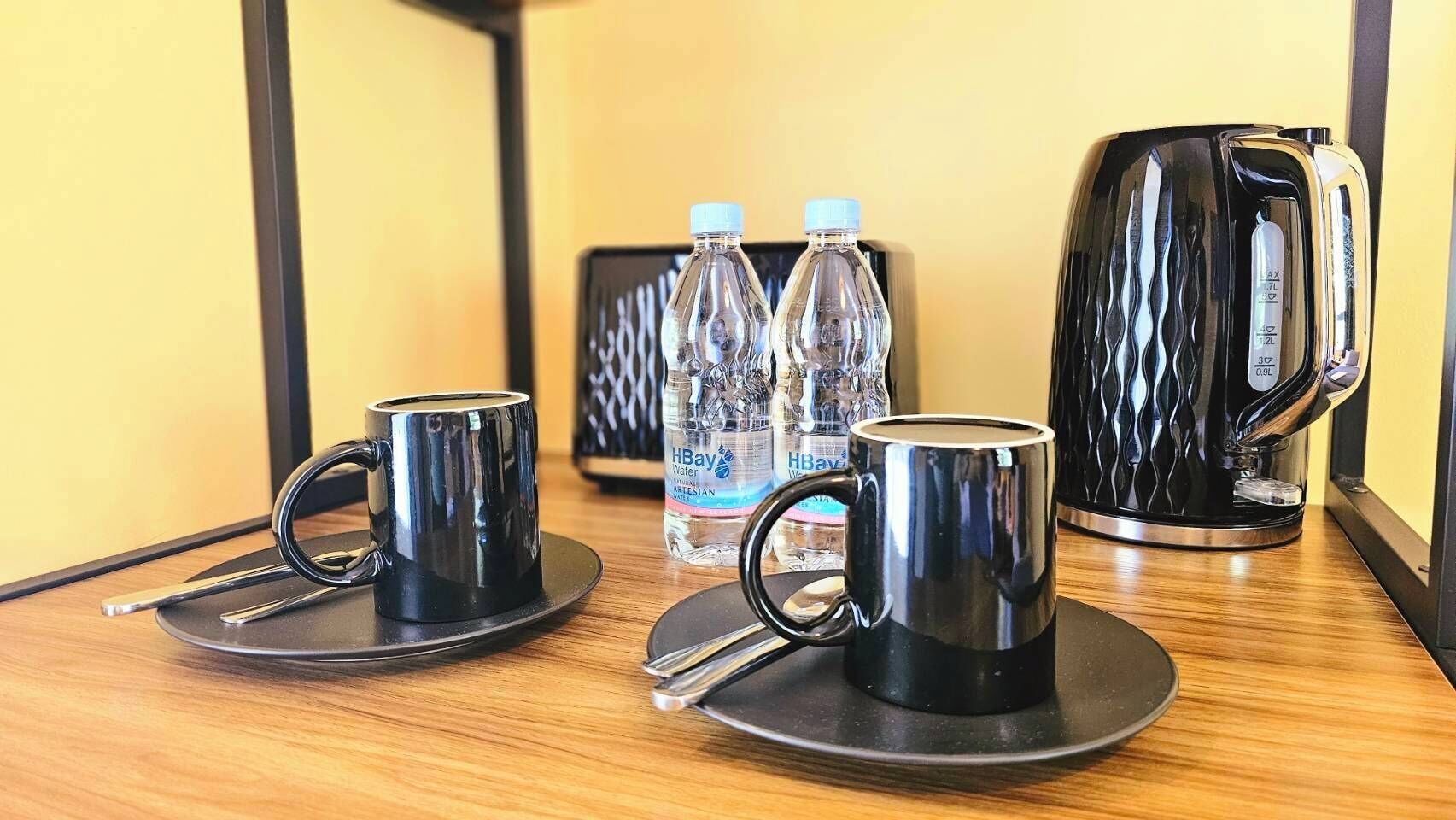 A hotel room with a wooden table, two black mugs, a kettle, and three water bottles.