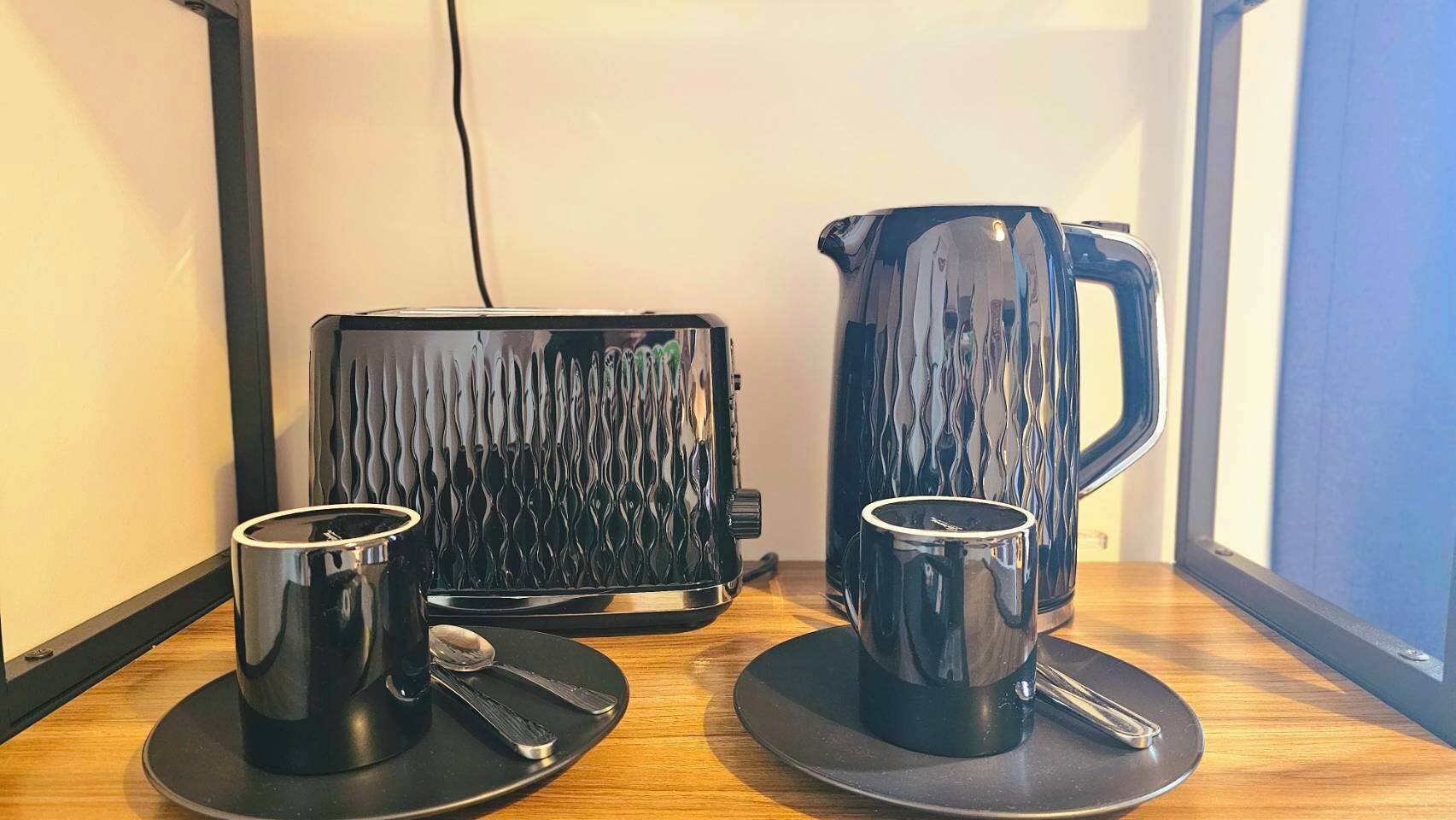 A room with a wooden table, two black mugs with saucers, a black tray, and a black electric kettle.