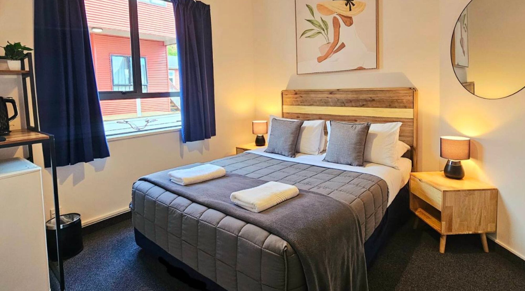 Un dormitorio ordenado con una cama grande, dos mesitas de noche con lámparas, una planta en la mesita de noche y una ventana con cortinas.