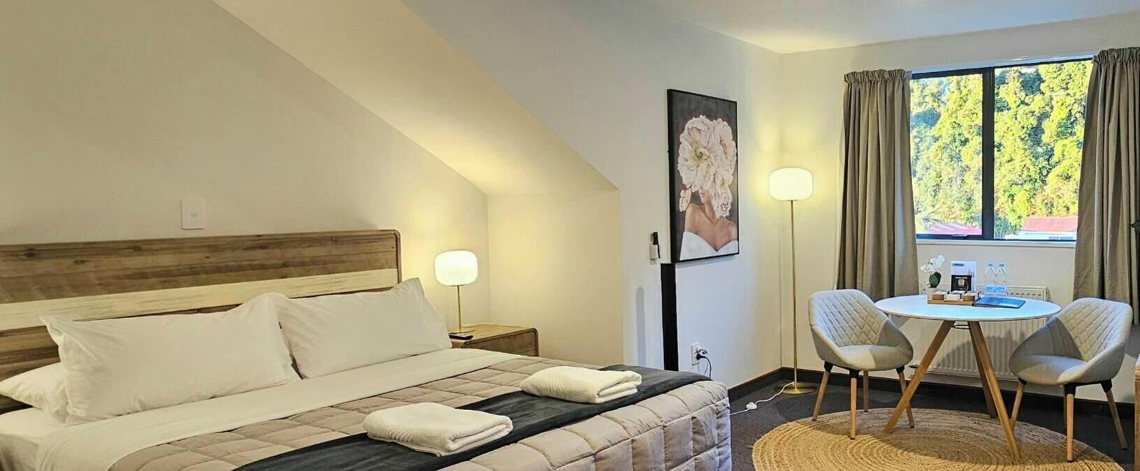 A hotel room with a bed, a nightstand, a bedside table, a lamp, a framed picture, a window with curtains, and a small round table with two chairs.