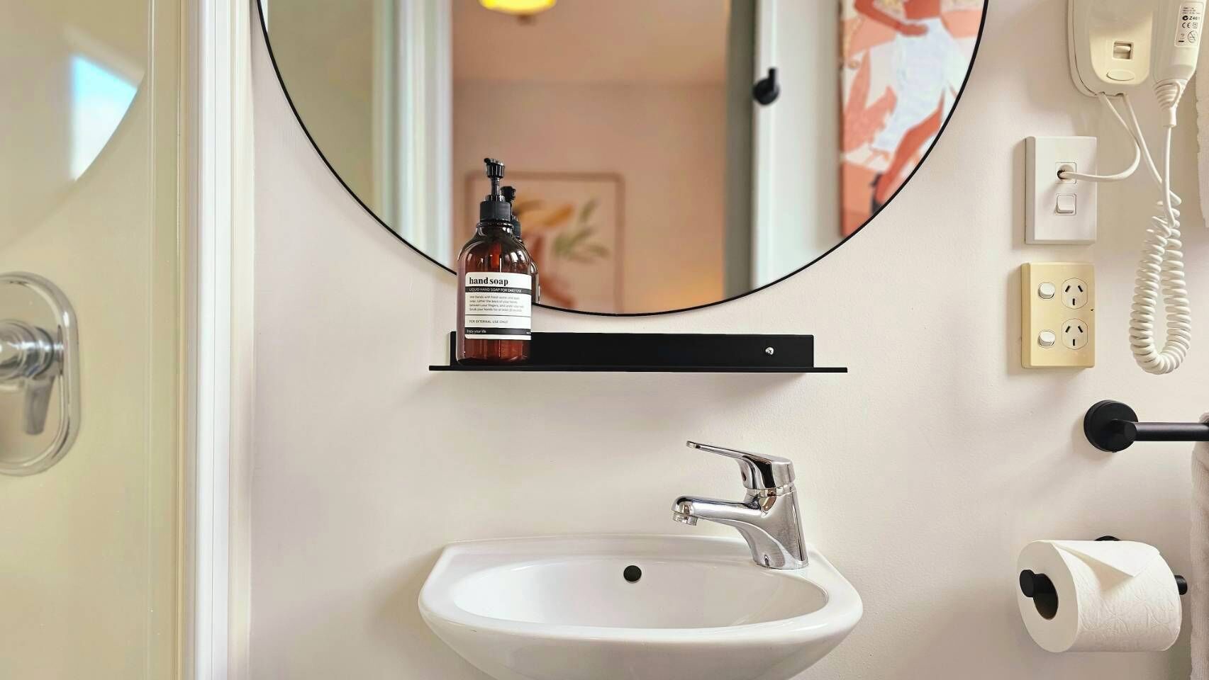 Une salle de bain avec un lavabo blanc, un miroir, un distributeur de savon liquide, un porte-rouleau de papier toilette et un téléphone fixé au mur.