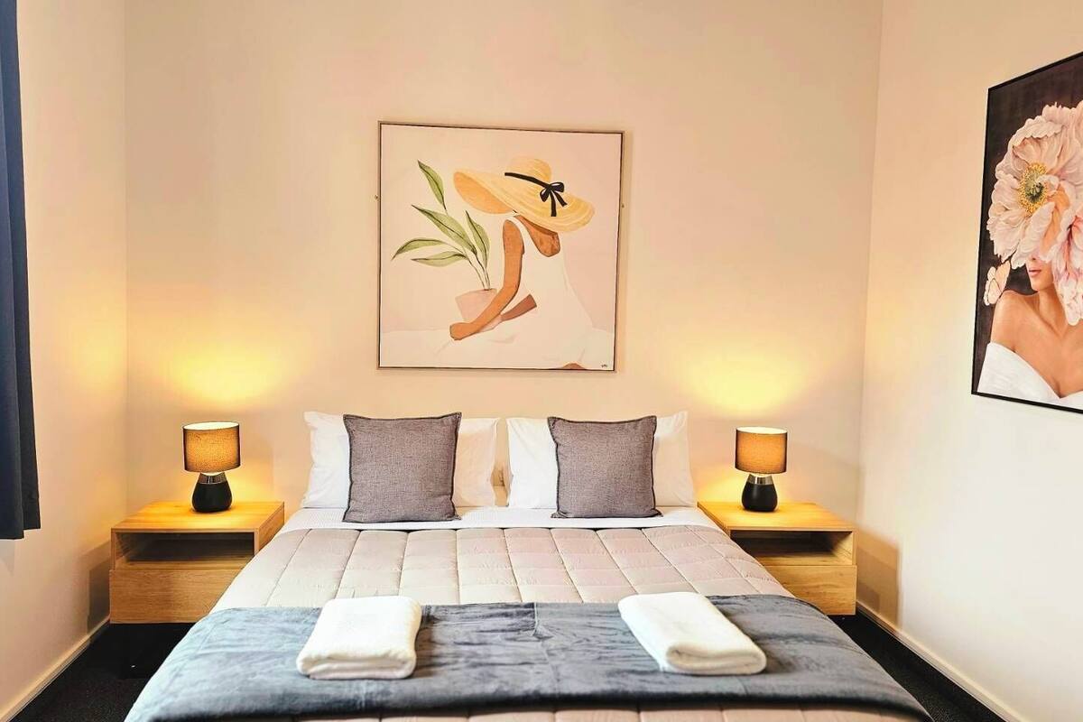 Une chambre d’hôtel comprenant un lit, deux tables de chevet, une commode, un tableau accroché au mur et une fenêtre avec des rideaux.