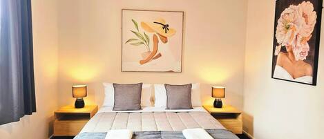 A hotel room with a bed, two bedside tables, a nightstand, a wall-mounted painting, and a window with curtains.