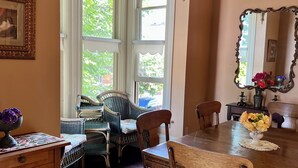 A dining area with a wooden table, chairs, a patterned rug, wicker furniture, and a large window with a view of greenery. - 2 bedroom Waterfront Victorian Apartment, with amazing views!  (Eureka)