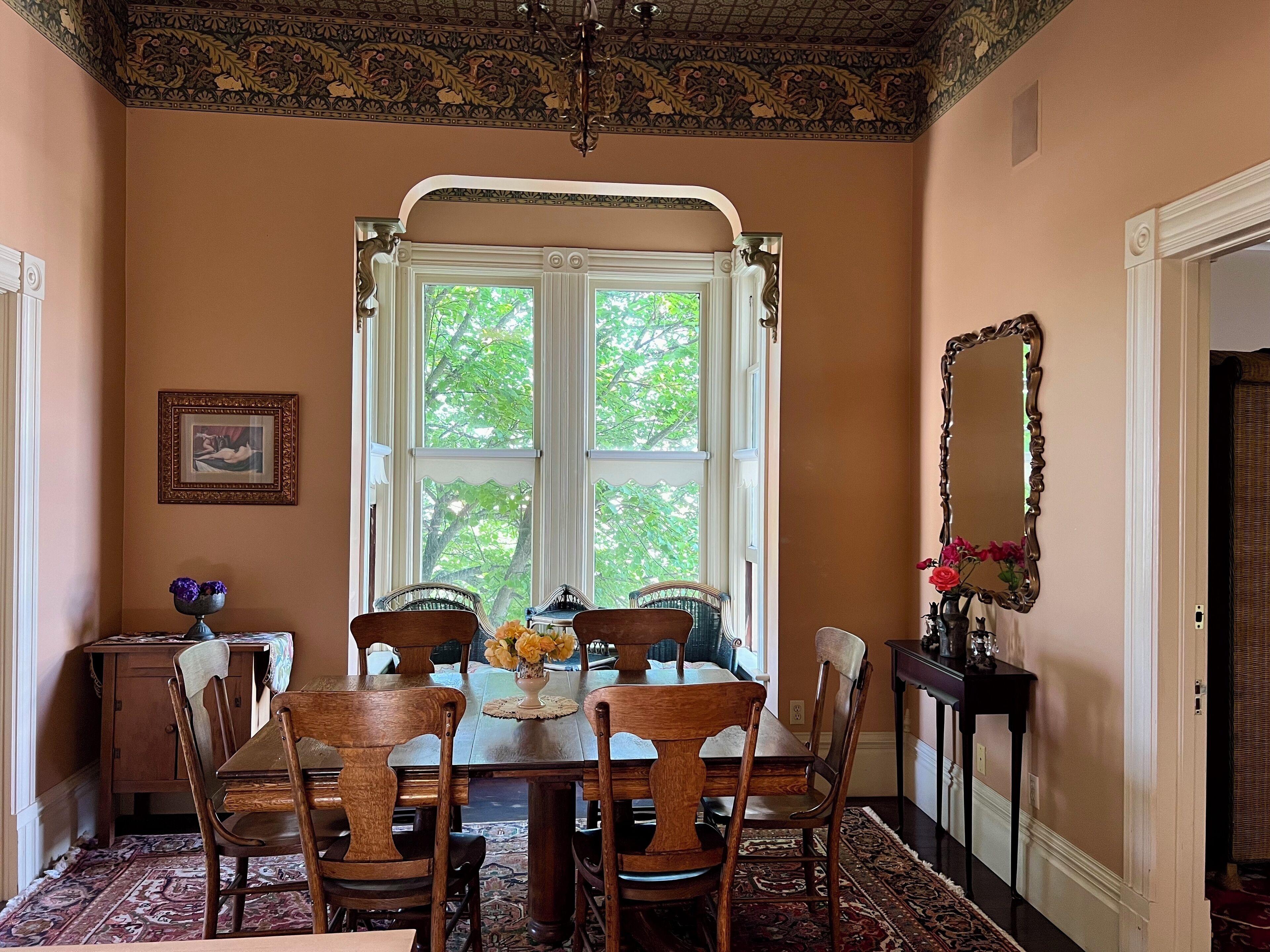 A dining room with a large window, ornate ceiling, and a mirror on the wall.