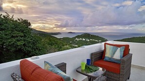 A balcony with wicker furniture and cushions, overlooking a scenic landscape with hills and the sea.