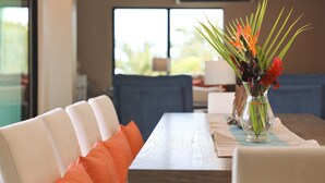 A dining table with white chairs and orange cushions, a vase with flowers, and a view of greenery outside.