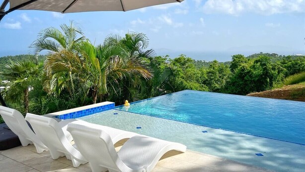 A swimming pool with white lounge chairs and a large umbrella, surrounded by lush greenery and a view of the ocean.