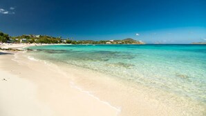 Una playa con agua turquesa cristalina, orilla de arena y edificios al fondo.