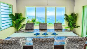 A dining area with a table set for four, surrounded by wicker chairs, with a view of a coastal landscape through large windows.