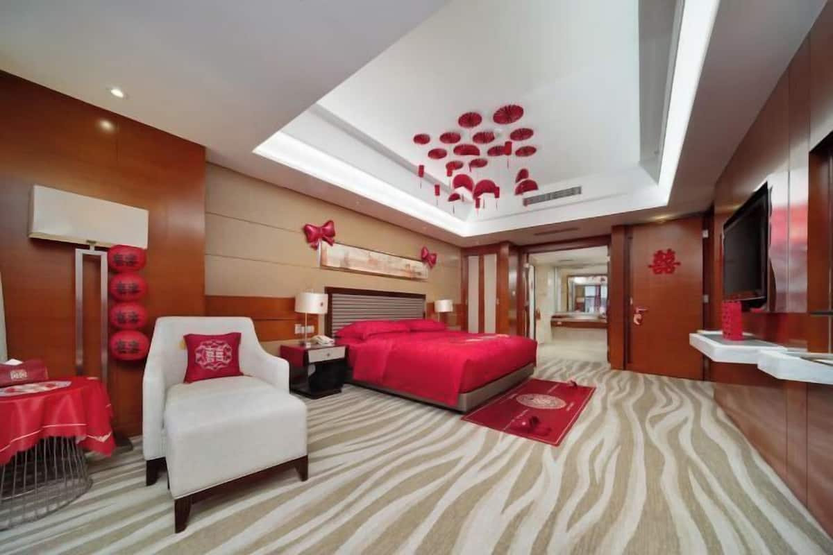 Una habitación de hotel con cama, silla, escritorio, televisión y techo con adornos rojos.
