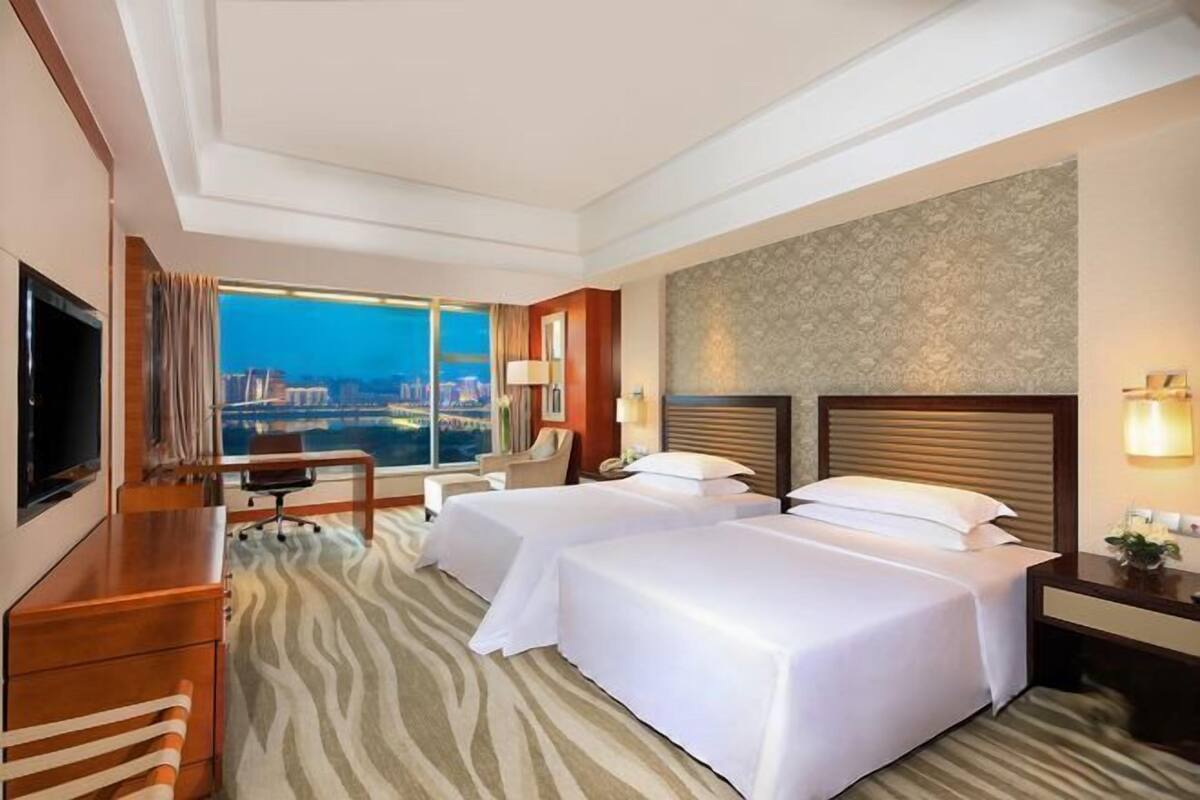 Habitación de hotel con dos camas, un escritorio, una silla, un televisor y un ventanal con vistas a la ciudad.